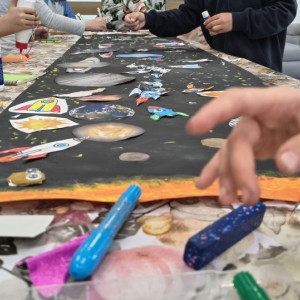 Dzieci tworzą na długim, czarnym papierze kosmiczną mozaikę z wyklejankami planet, rakiet i gwiazd. Na pierwszym planie widać kolorowe kredki i rękę dziecka.