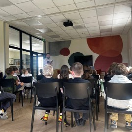 Grupa dzieci siedzi na krzesłach, słuchając mężczyzny czytającego książkę. W tle znajduje się kolorowa ściana z geometrycznymi kształtami.