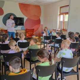 Mężczyzna w kolorowej koszuli prowadzi zajęcia dla grupy dzieci siedzących na krzesłach w sali z kolorowym muralem na ścianie.