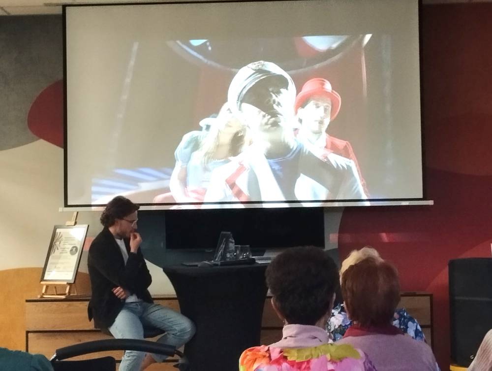 Ekran projekcyjny z fragmentem filmu lub prezentacji, na kt&oacute;rym widoczni są ludzie w strojach, być może cyrkowych. Na pierwszym planie siedzący mężczyzna w okularach, zamyślony.