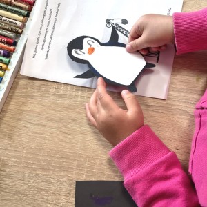 Dziecko w r&oacute;żowej bluzce przykleja wyciętego z papieru pingwina na kartkę z książki. Obok leży pudełko z kredkami.