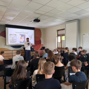 Prelegent z brodą, w białej koszuli, stoi przed ekranem z napisem 