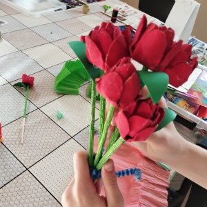 Zrobione z papieru czerwone r&oacute;że z zielonymi liśćmi i łodyżkami, trzymane w dłoniach. W tle widać st&oacute;ł z materiałami do rękodzieła.