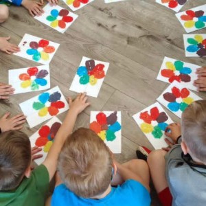 Dzieci siedzą na podłodze w kręgu, trzymając w rękach kartki z kolorowymi malunkami przypominającymi kwiaty. Każdy obrazek przedstawia centralny punkt i otaczające go płatki w r&oacute;żnych barwach.