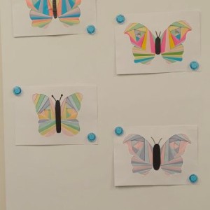 Cztery kolorowe, geometryczne motyle namalowane na białych kartkach, przyczepione do białej ściany niebieskimi magnesami. Motyle mają tęczowe skrzydła złożone z wielu tr&oacute;jkąt&oacute;w i prostokąt&oacute;w.