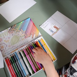 Dłoń dziecka wybiera kredkę z otwartego pudełka pełnego kolorowych kredek. Obok leży szkicownik z rysunkiem i plansza z ramkami do komiksu.